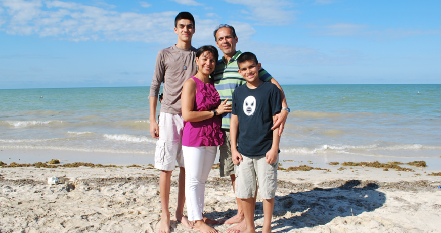 Family of four standing and smiling on a sunny beach with blue sky and ocean in the background.